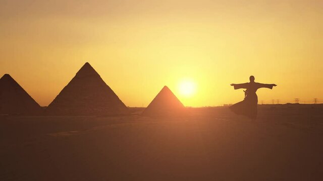 Silhouette of woman in traditional arab dress rises her arms at the sunset with pyramids on the background.