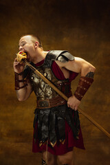 Portrait of young man, medieval warrior or knight in war equipment eating burger isolated on vintage dark background. Comparison of eras, art, history, humor, retro style concept