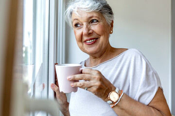 Close up profile view of old aged woman standing with pink mug looking out window. Senior woman having her coffee time at home. Happy elderly woman drinking a tea. Beautiful old lady behind the window