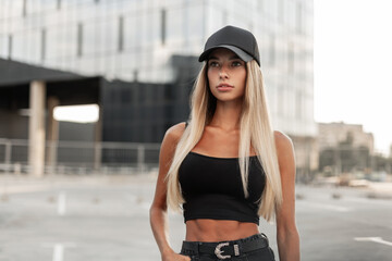 Pretty model hipster woman in a black baseball cap with a black stylish tank top and denim shorts with a belt stands in the city. Modern urban female style look outfit - obrazy, fototapety, plakaty