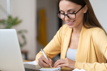 Young student woman studying from home use laptop computer watch educational video or e-learning courses making notes in notebook. Female entrepreneur passing certificate training learning online