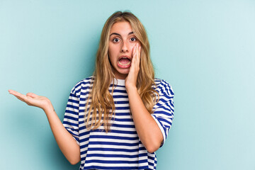 Young caucasian blonde woman isolated on blue background  impressed holding copy space on palm.