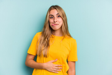 Young caucasian blonde woman isolated on blue background  touches tummy, smiles gently, eating and satisfaction concept.