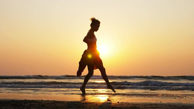 Silhouette of young girl dancing at sunset. Female artist is dancing on the sandy beach against orange sunset, slow motion