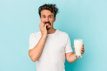 Young caucasian man holding a glass of milk isolated on blue background biting fingernails, nervous and very anxious.