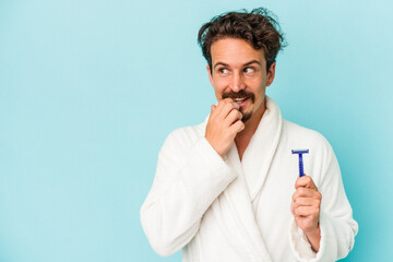 Young caucasian man holding a razor blade isolated on blue background relaxed thinking about something looking at a copy space.