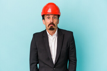 Young architect caucasian man isolated on blue background shrugs shoulders and open eyes confused.