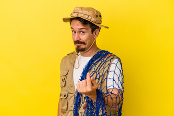 Young caucasian fisherman holding net isolated on yellow background pointing with finger at you as if inviting come closer.