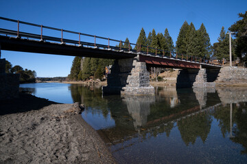 The bridge across the rive connecting two lakes. Its reflection in the water surface. The beach shore and forest in a sunny summer day with a clear blue sky. 