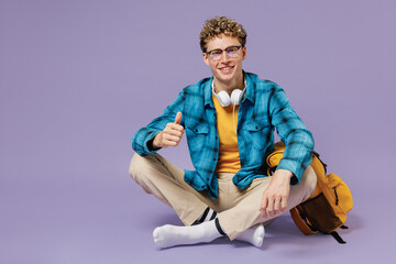 Full body young boy teen student wear casual clothes backpack headphones glasses sit crossed legs isolated on violet background studio portrait. Education in high school university college concept