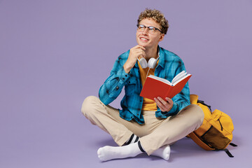 Fototapeta na wymiar Full size young boy teen student wear casual clothes backpack headphones glasses sit reading book isolated on violet background studio portrait. Education in high school university college concept