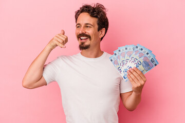 Young caucasian man holding banknotes isolated on pink background showing a mobile phone call gesture with fingers.