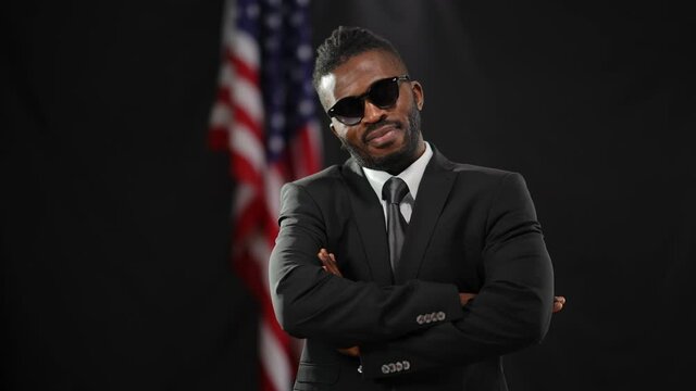 Proud confident African American man putting on sunglasses crossing hands in camera flashes as dollars falling. Portrait of successful elections winner posing at black background with national flag