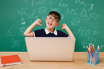 Young happy male kid school boy 5-6 year old in t-shirt glasses sit use laptop pc cocmputer point finger on himself isolated on green wall chalk blackboard background Childhood children kids concept