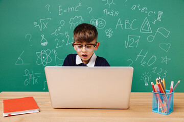 Young smart male kid school boy 5-6 year old in t-shirt glasses sit using laptop pc cocmputer isolated on green wall chalk blackboard background Childhood children kids education lifestyle concept