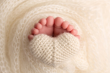 The feet of a newborn baby are wrapped in a knitted blanket. The fingers of a newborn baby are holding a white knitted heart.