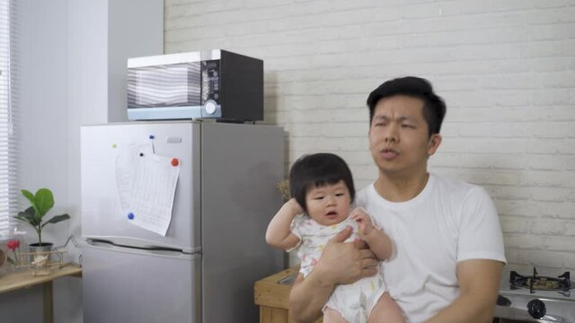 hyped asian dad is singing air karaoke into his fist while his sleepy baby girl is pulling hair in his arm at home kitchen during daytime.