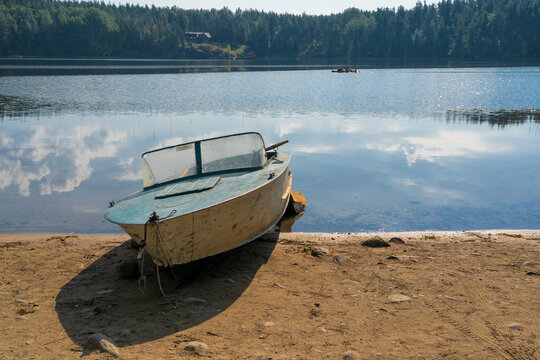 An old metal motor boat lies on the shore of the lake. You can see the opposite shore with a forest and a reflection of the sky in the water.