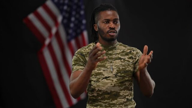 Handsome bearded African American lieutenant sergeant persuading recruits with USA flag at black background. Portrait of confident brave patriotic man talking and gesturing. Military service concept