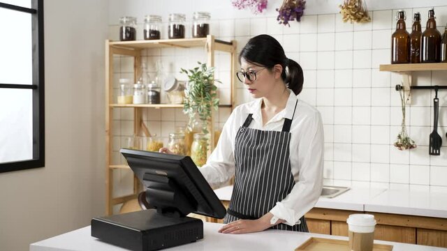 female asian coffee restaurant manager wearing an apron is making inventory checkup on the checkout counter computer with concentration.