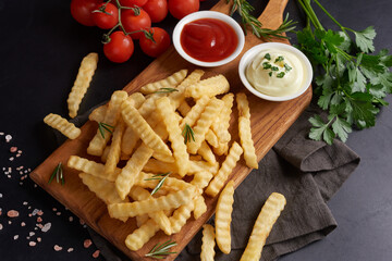 Homemade baked potato fries with mayonnaise, tomato sauce and rosemary on wooden board. tasty french fries on cutting board, in brown paper bag on black stone table background, unhealthy food.
