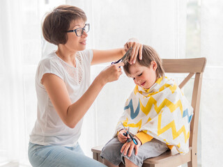 Obraz na płótnie Canvas Mother cuts her son's hair by herself. Little boy sits, covered with cloth, and holds pair of scissors. New normal in case of coronavirus COVID-19 quarantine. Beauty at home lifestyle.
