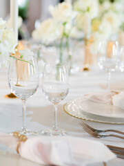 Table serves for banquet. Transparent wine glasses, plates with napkins and shiny cutlery on white table cloth at sunlight.