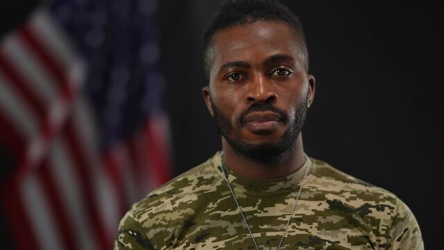 Unshaved confident serious African American soldier looking away and turning to camera. Portrait of brave military man posing at black background with USA flag. Confidence and patriotism