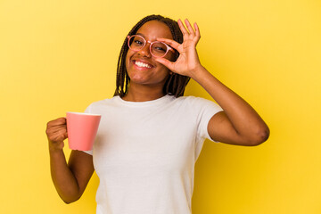 Young african american woman holding a mug isolated on yellow background  excited keeping ok gesture on eye.