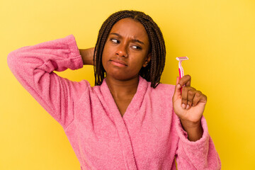 Young african american woman wearing a bathrobe holding a razor blade isolated on yellow background  touching back of head, thinking and making a choice.