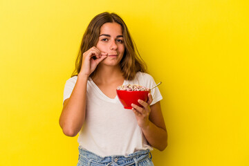 Young caucasian woman holding cereals isolated on yellow background  with fingers on lips keeping a secret.