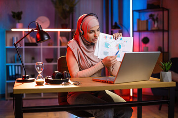 Muslim woman in headset using wireless laptop at home for video call. Young female in hijab showing financial report during online meeting. Concept of people and freelance.