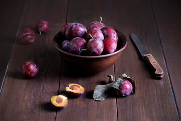 Still life with fresh ripe plums in a clay ceramic bowl and a knife on a dark wooden surface....