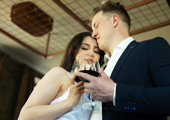 The groom in a suit and the bride in a wedding dress hold glasses of red wine drinking.