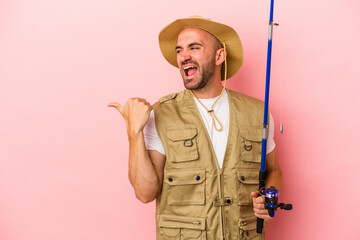 Young bald fisherman holding a rod isolated on pink background  points with thumb finger away, laughing and carefree.