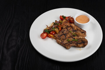 Fried pork chop with grilled vegetables and sauce on white plate on black wooden background