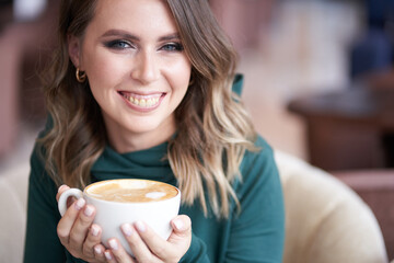 A happy beautiful brunette with a beautiful manicure, holding a cup of coffee in her hands, wearing a green turtleneck, sitting in a cafe, looks directly into the camera. High quality photo