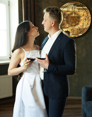 The groom in a suit and the bride in a wedding dress hold glasses of red wine drinking.