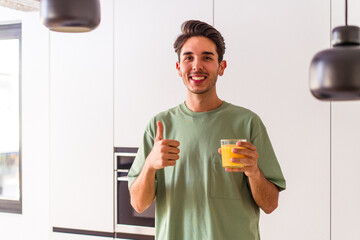 Young mixed race man drinking orange juice in his kitchen smiling and raising thumb up