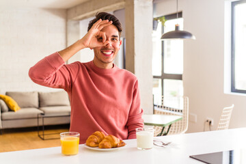 Fototapeta na wymiar Young mixed race man having breakfast in a kitchen on the morning excited keeping ok gesture on eye.