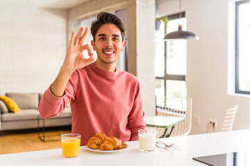 Young mixed race man having breakfast in a kitchen on the morning cheerful and confident showing ok...