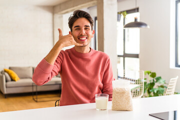 Young mixed race man eating oatmeal and milk for breakfast in his kitchen showing a mobile phone call gesture with fingers.