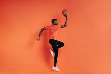 Fototapeta na wymiar Side view of sportsman jumping in the air with basket ball in hand. Male in sportswear exercising indoors at an orange wall.
