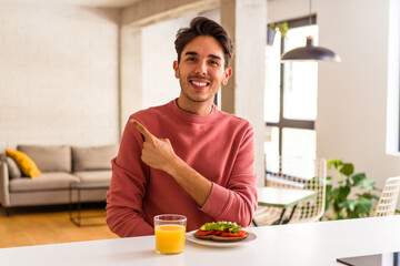 Fototapeta na wymiar Young mixed race man having breakfast in his kitchen smiling and pointing aside, showing something at blank space.