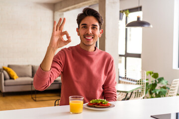 Fototapeta na wymiar Young mixed race man having breakfast in his kitchen cheerful and confident showing ok gesture.