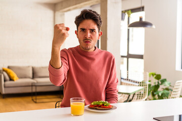 Fototapeta na wymiar Young mixed race man having breakfast in his kitchen showing fist to camera, aggressive facial expression.