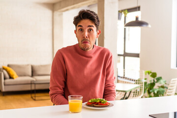 Fototapeta na wymiar Young mixed race man having breakfast in his kitchen shrugs shoulders and open eyes confused.