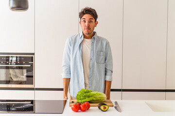 Young mixed race man preparing a salad for lunch shrugs shoulders and open eyes confused.