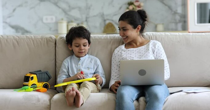 Indian mom put laptop on laps talks to little adorable son sit nearby drawing on magnetic erasable board. Family spend free time together, enjoy communication. Hobby, weekend leisure activity concept