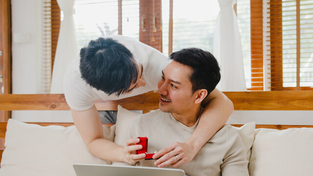 Young Asia gay couple propose at modern home, LGBTQ men happy smiling have romantic time while proposing and marriage surprise wear wedding ring in living room at house. Anniversary romantic concept.